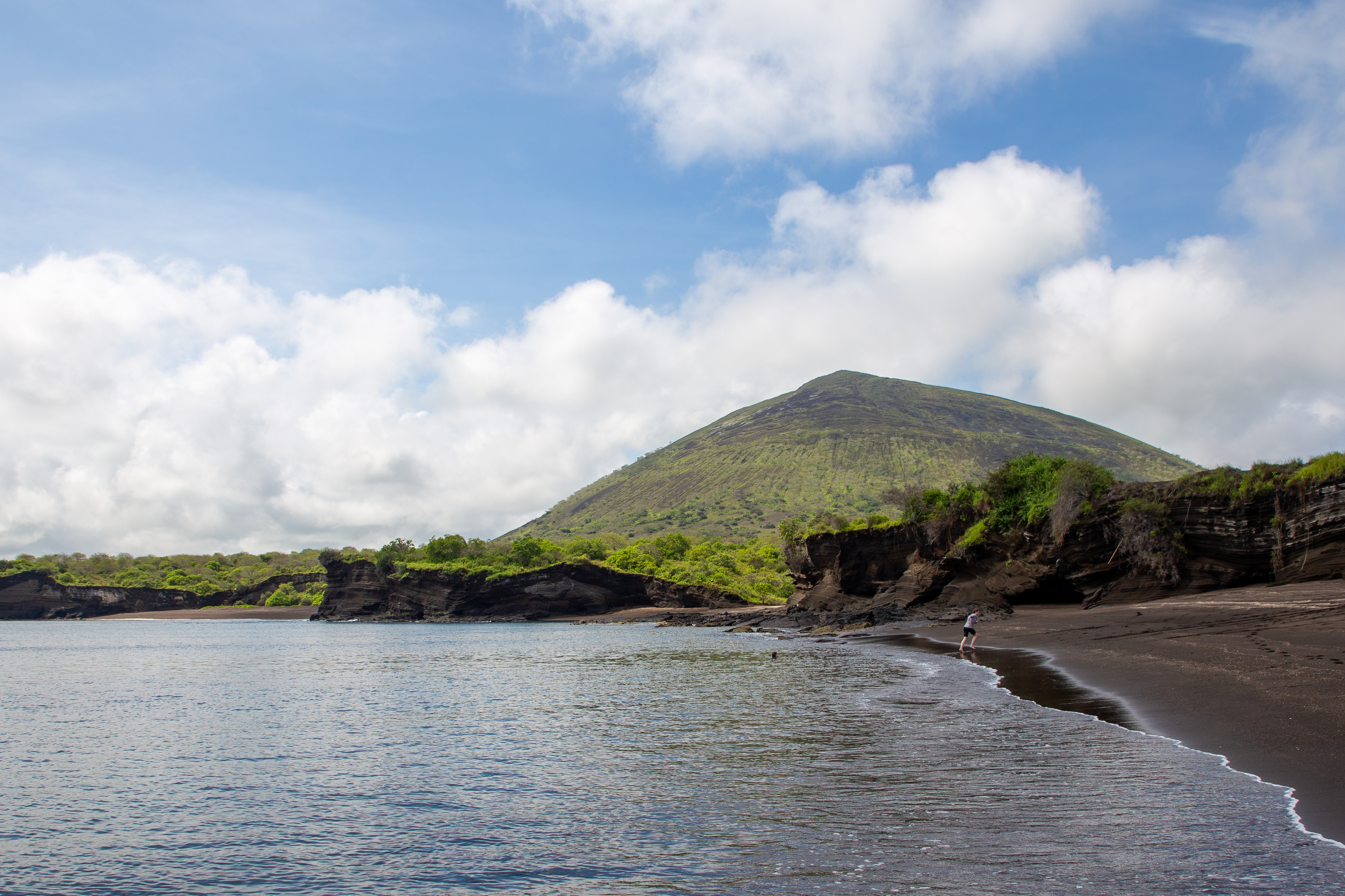 Black sand beach curving toward a green volcanic cone under a cloudy sky